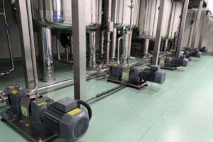 Fila de bombas industriales instaladas en un suelo limpio y verde, conectadas a tuberías de acero inoxidable en una instalación moderna.